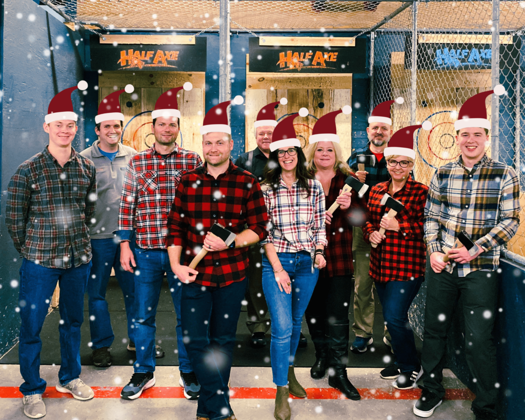 A photo of a group of adult men & women in flannel shirts with santa hats & snow falling in front of axe throwing targets