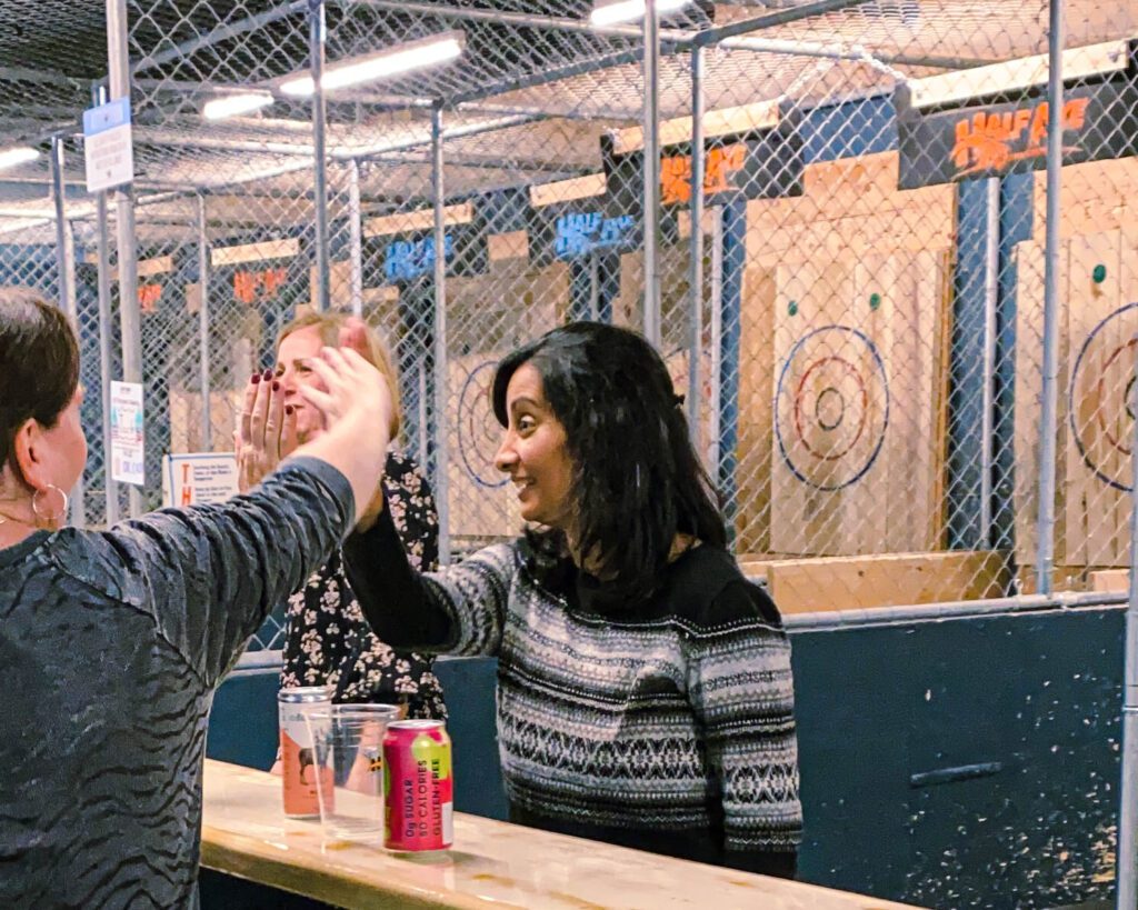 Two women high-fiving two other women on the opposite side of a counter. Wooden targets are behind them. 