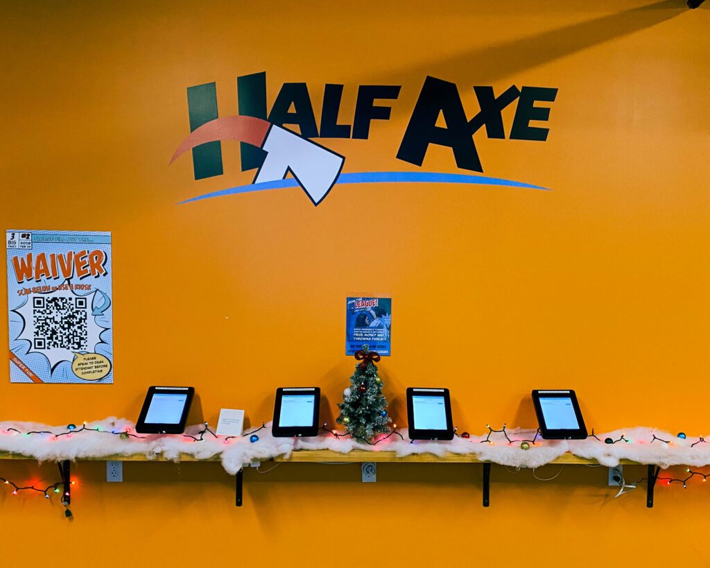 A large orange wall with big text saying, "Half Axe", with a shelf of 5 ipads beneath the text. On this shelf is fake snow and holiday decorations. 